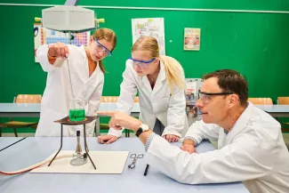 Twee leerlingen en een docent in labjassen doen een experiment met de gasbrander.